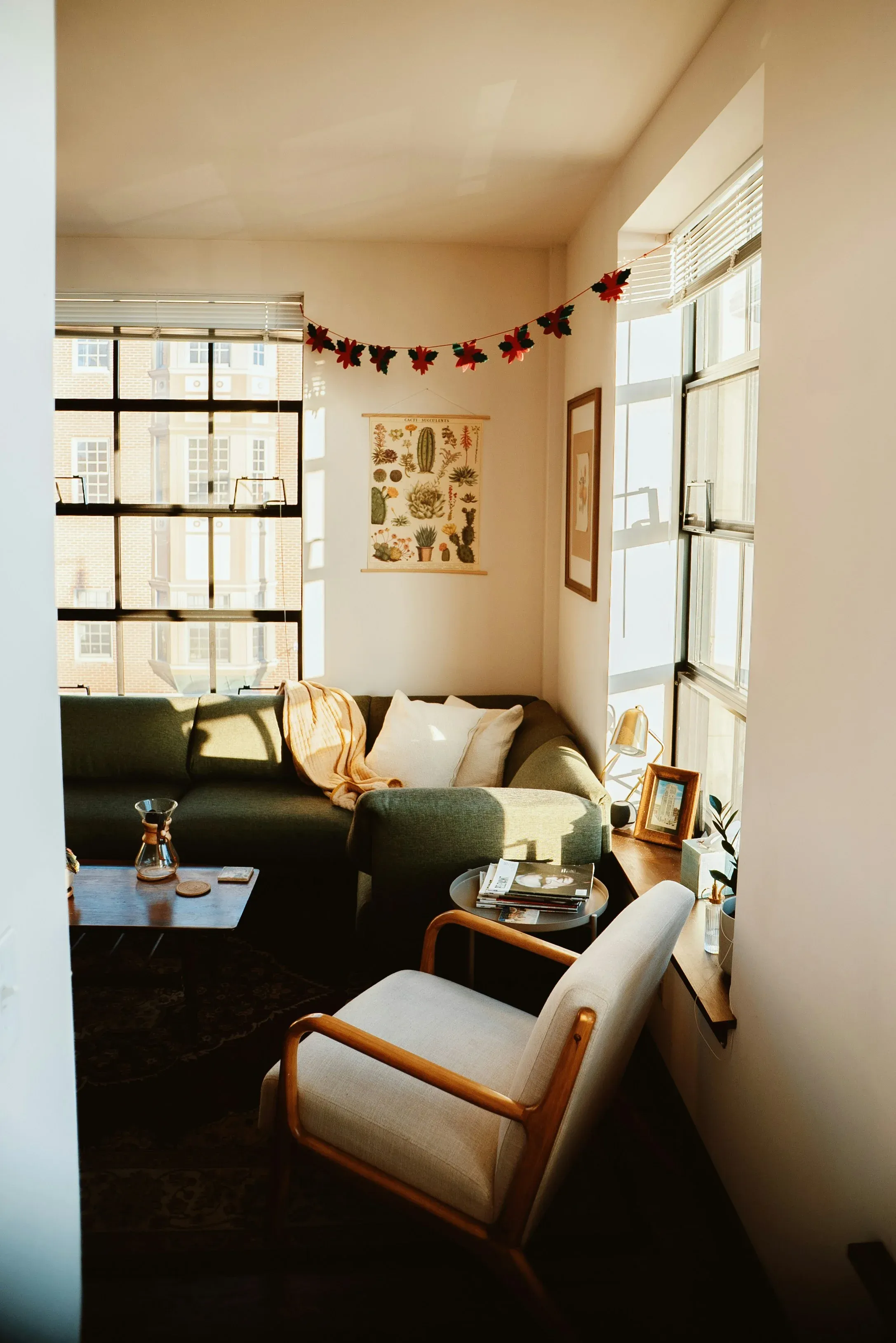 A warmly lit sitting area with a green sofa, framed artwork, and corner windows